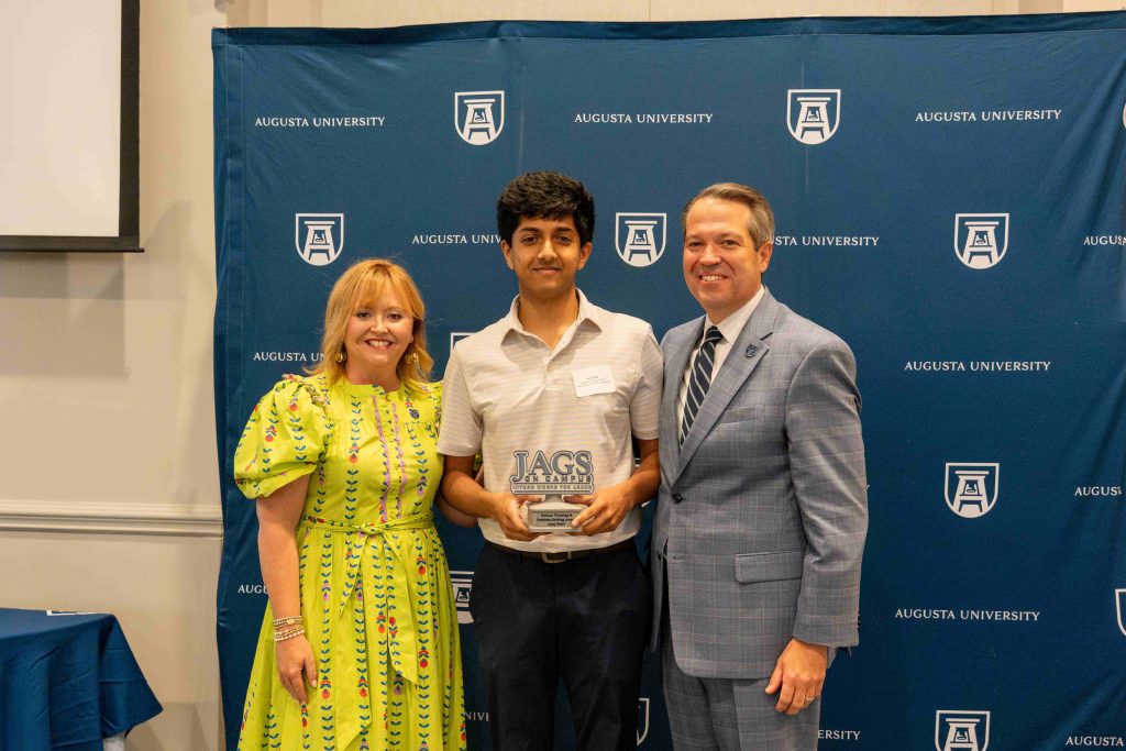 A college-aged boy holds an award, flanked by an older man on one side and an older woman on the other.