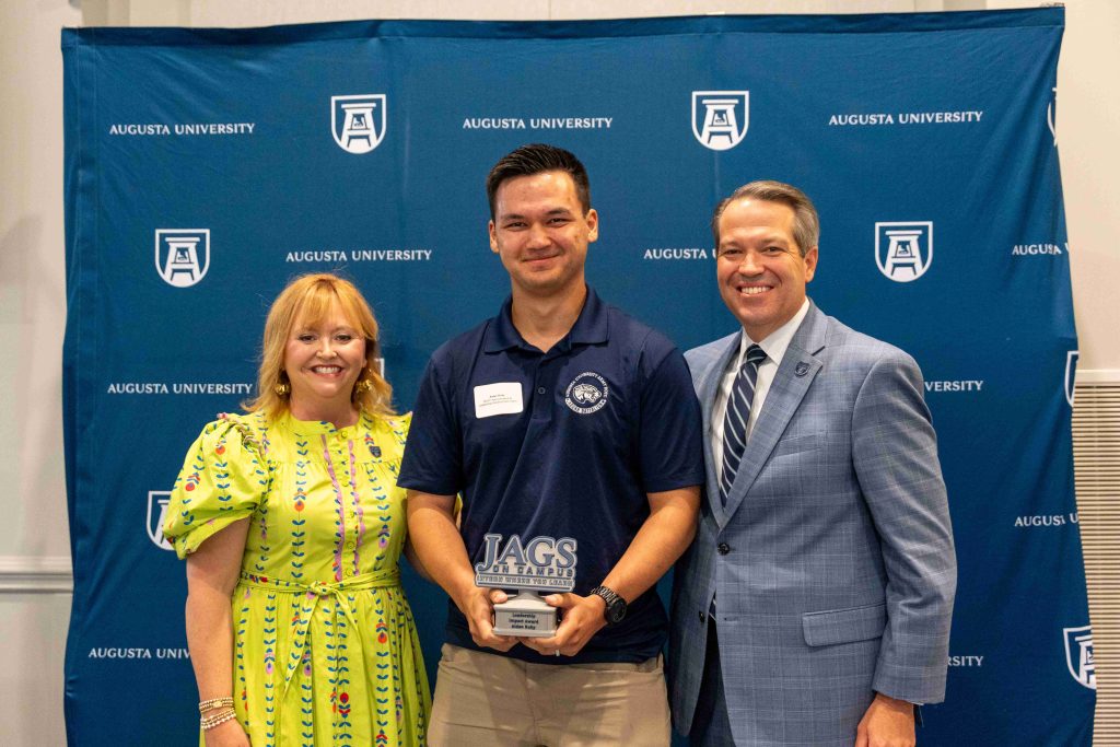 A college-aged boy holds an award, flanked by an older man on one side and an older woman on the other.