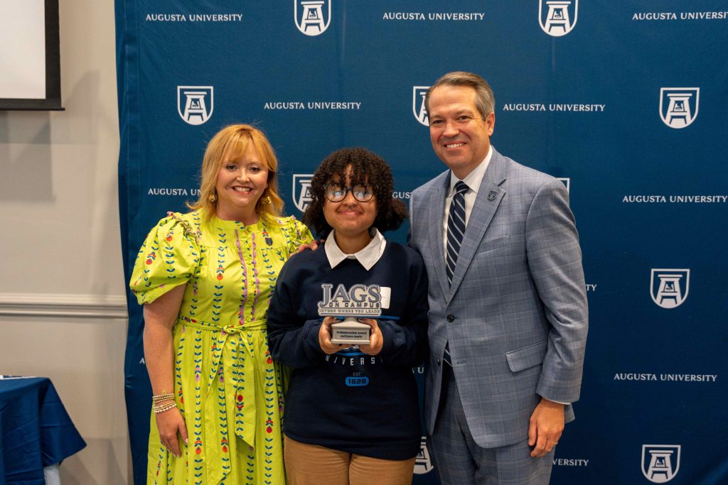A college-aged girl holds an award, flanked by an older man on one side and an older woman on the other.