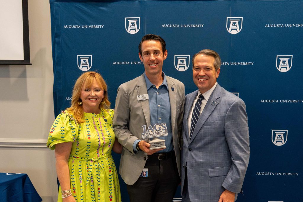 A man holds an award, flanked by an older man on one side and an older woman on the other.