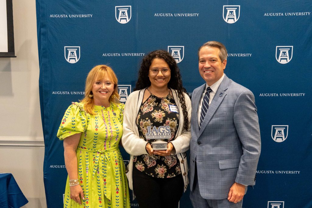A college-aged girl holds an award, flanked by an older man on one side and an older woman on the other.