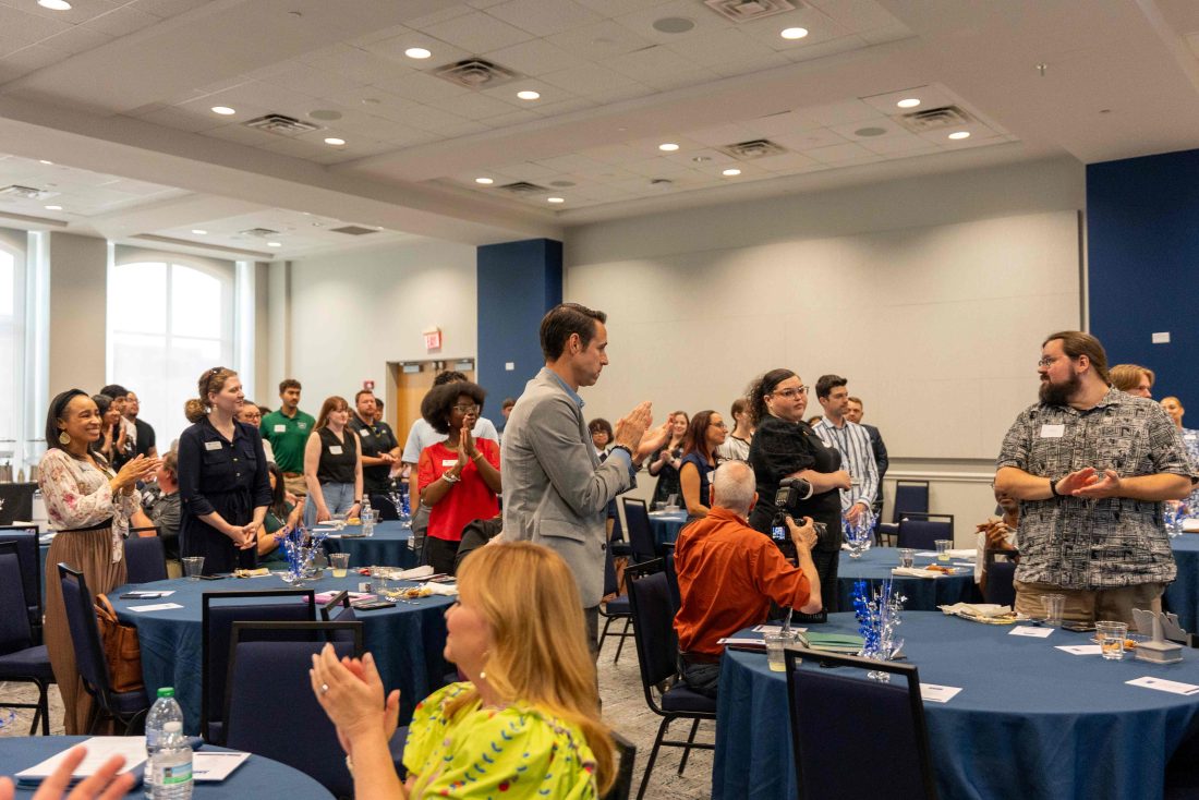 Men and women clapping while standing in between several tables.
