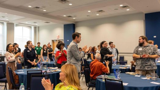 Men and women clapping while standing in between several tables.