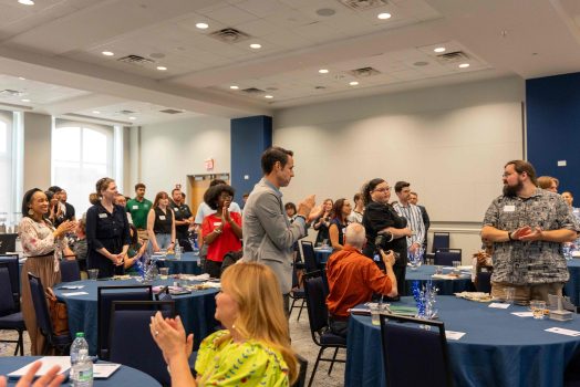 Men and women clapping while standing in between several tables.