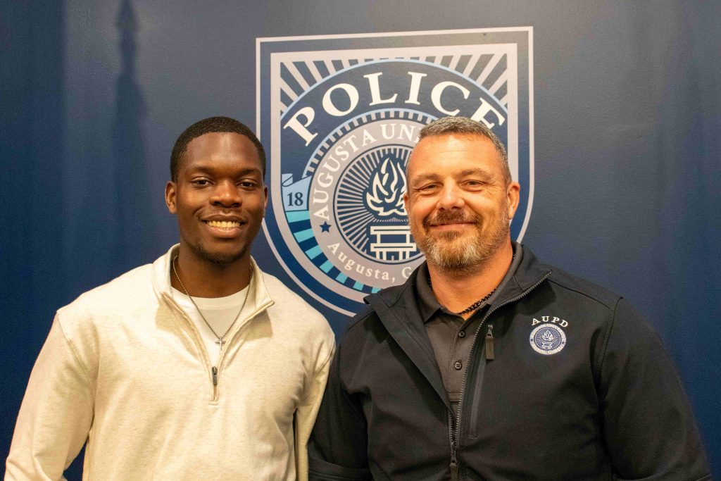 A young college-aged man and an older man smile for a photo in front of a police logo.