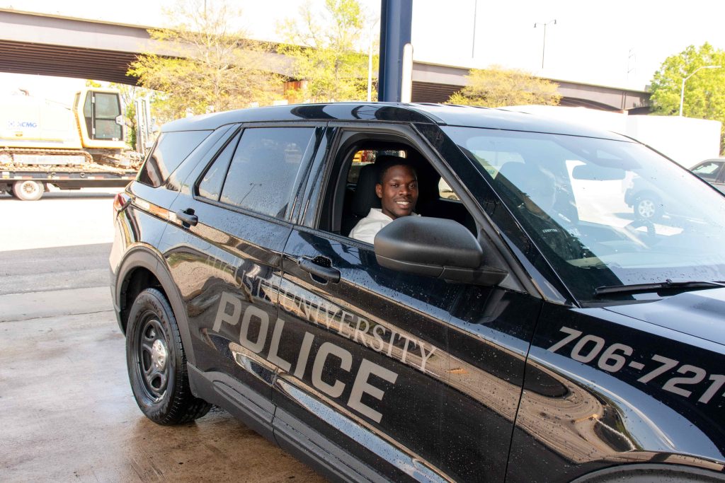 A young college-aged boy sits in the passenger seat of an Augusta University police car.