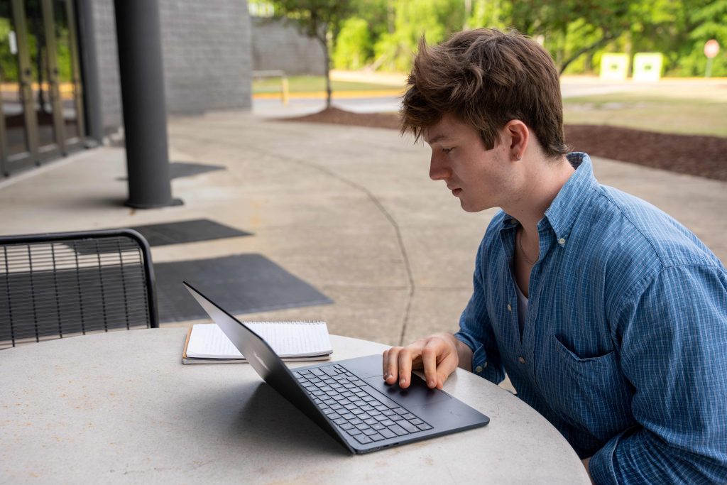 A college-aged boy sitting at an outdoor table scrolling on his laptop.