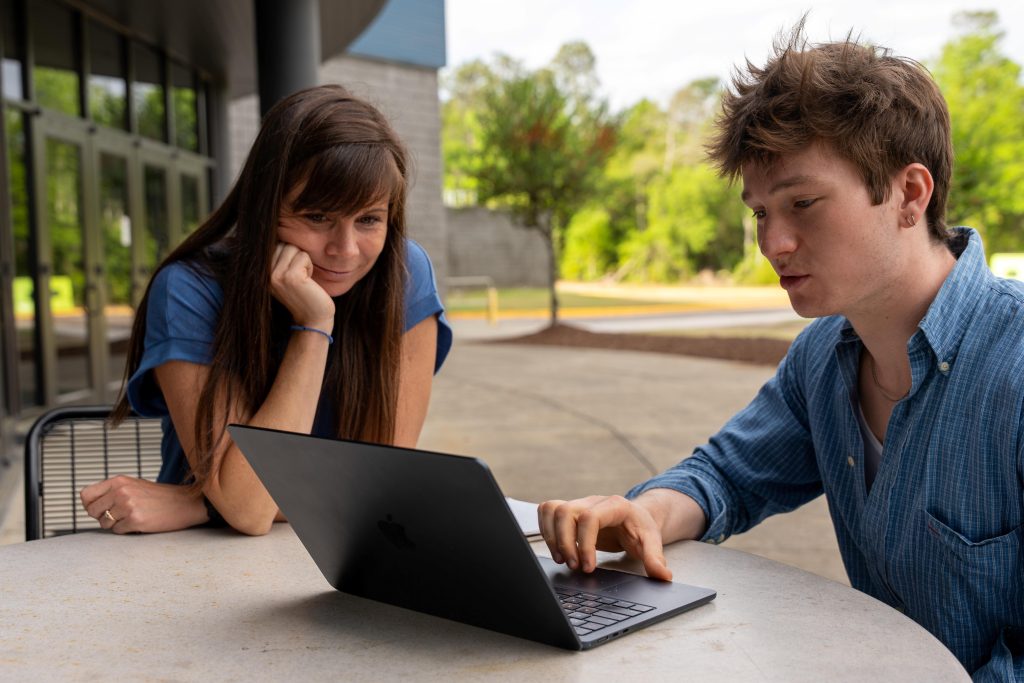 A college-aged boy and older woman sitting at an outdoor table looking at a laptop screen.