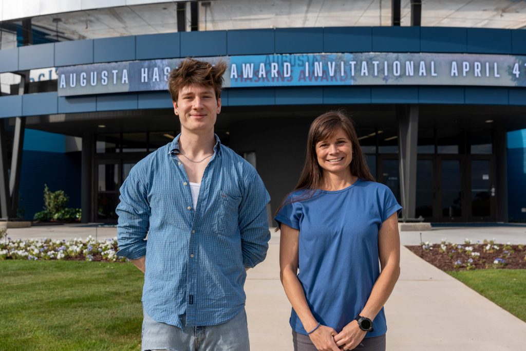 A college-aged boy and an older woman stand beside each other smiling, with a large building in the background.