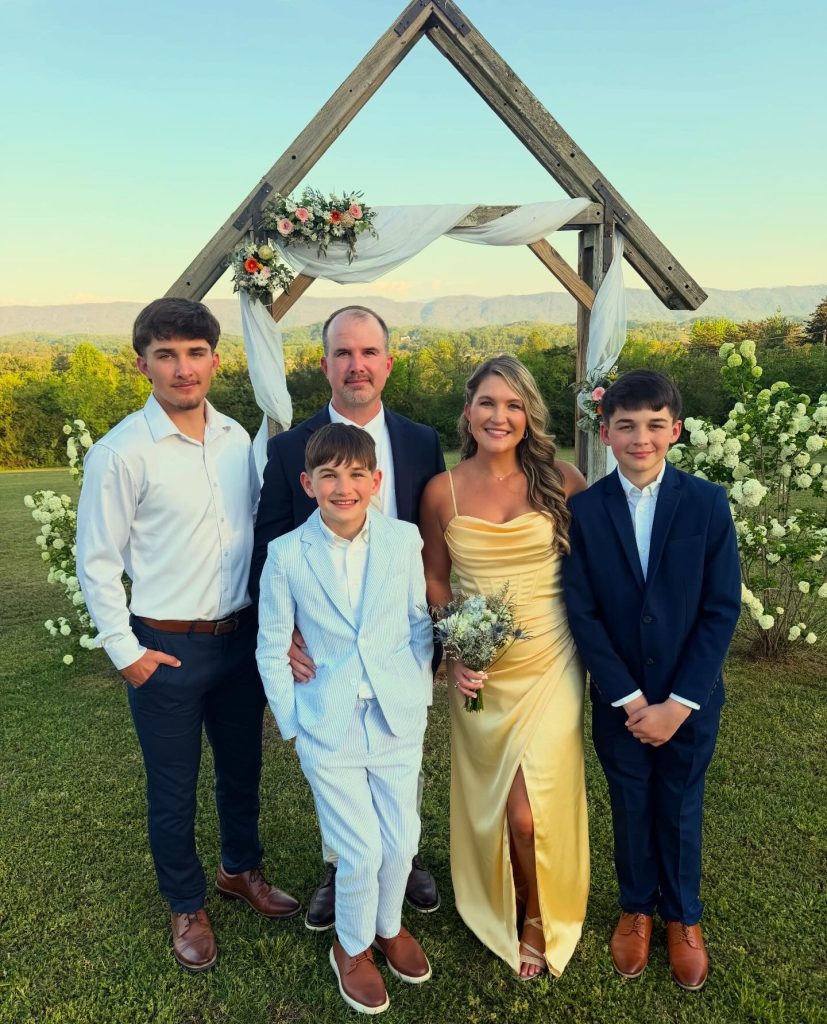 A husband and wife in wedding guest attire pose with their three sons in front of a wooden arch