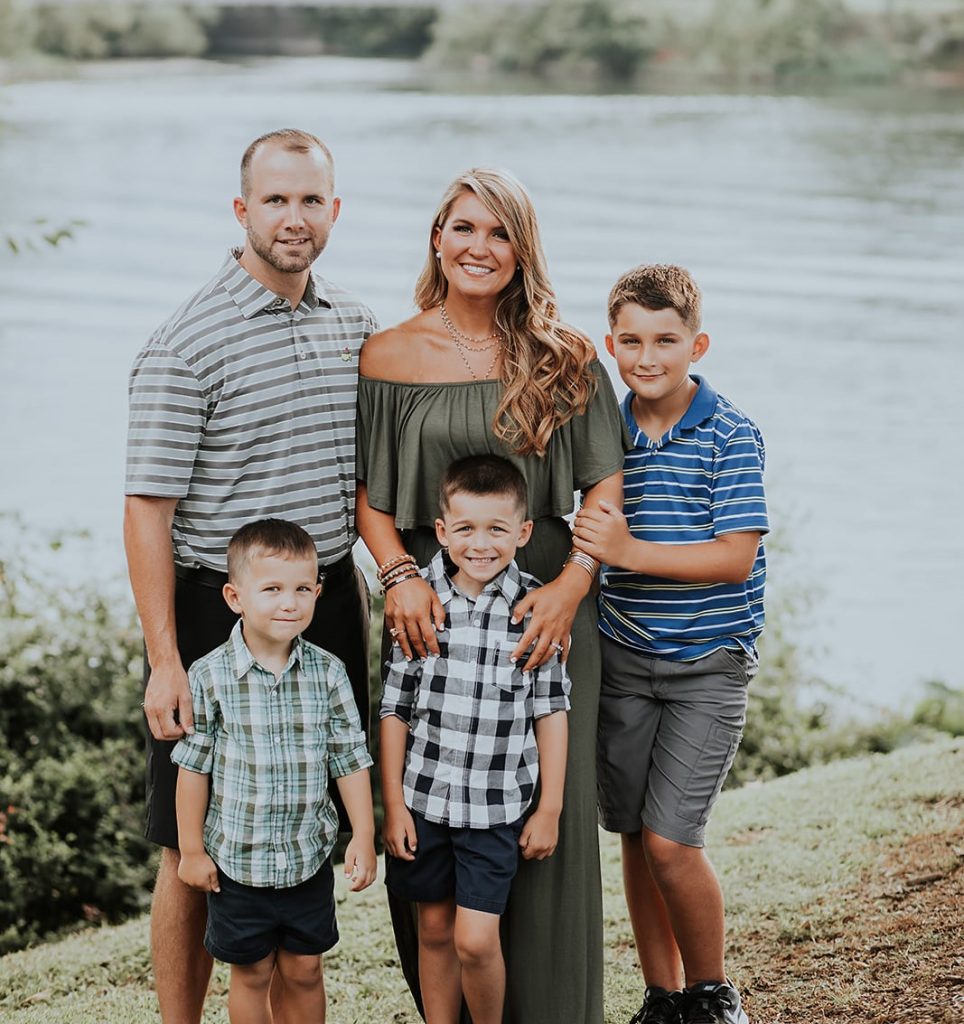 A husband and wife pose with their three young sons in front of a picturesque lake