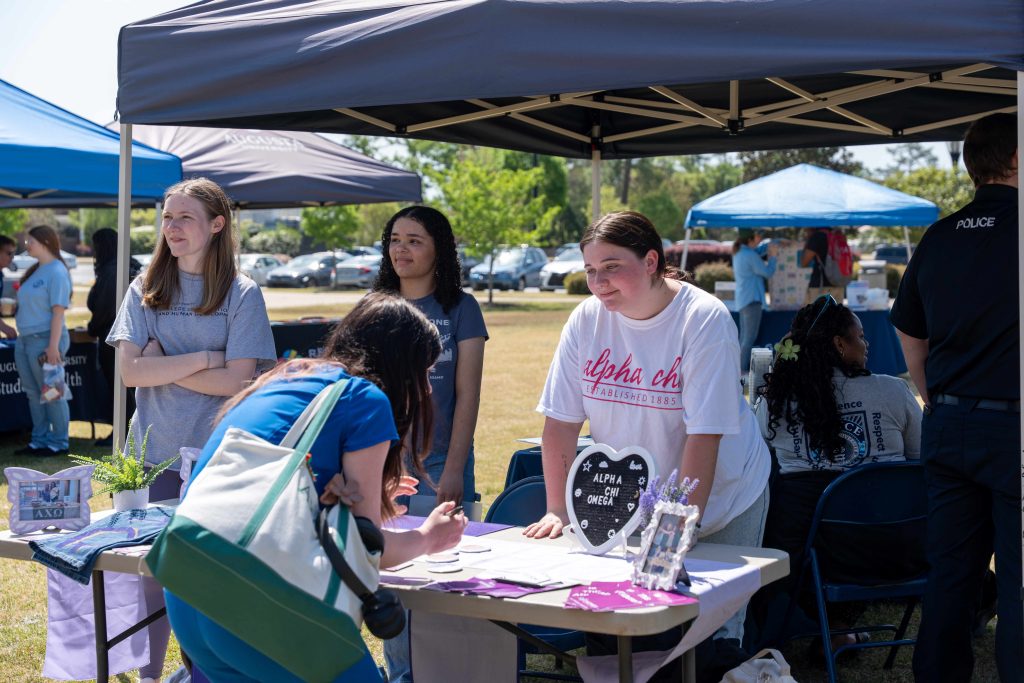At an outdoor tabling event, a college-aged woman leans down to write on a piece of paper while a tabling assistant speaks with her.