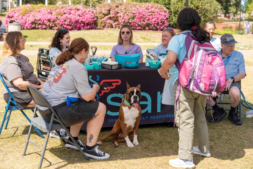 Several men and women sit around a table while a college-aged woman stands in front of it with a dog sitting beside her.