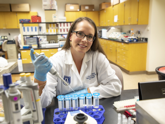 A woman wearing a lab coat holding a syringe smiles at the camera while sitting in a laboratory.