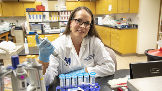 A woman wearing a lab coat holding a syringe smiles at the camera while sitting in a laboratory.