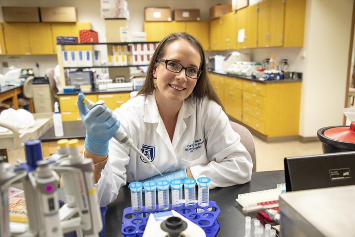 A woman wearing a lab coat holding a syringe smiles at the camera while sitting in a laboratory.
