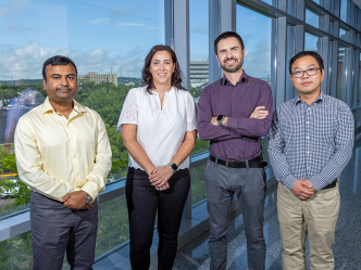 A group of four scientists - including three men and one woman - stand in a hallway of a modern research building looking out over a large hospital.