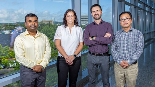 A group of four scientists - including three men and one woman - stand in a hallway of a modern research building looking out over a large hospital.