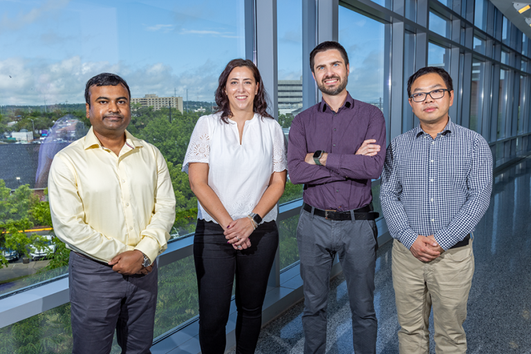 A group of four scientists - including three men and one woman - stand in a hallway of a modern research building looking out over a large hospital.