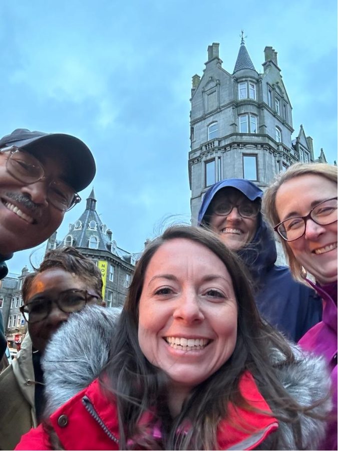 A group of men and women take a selfie with the background of buildings behind them.