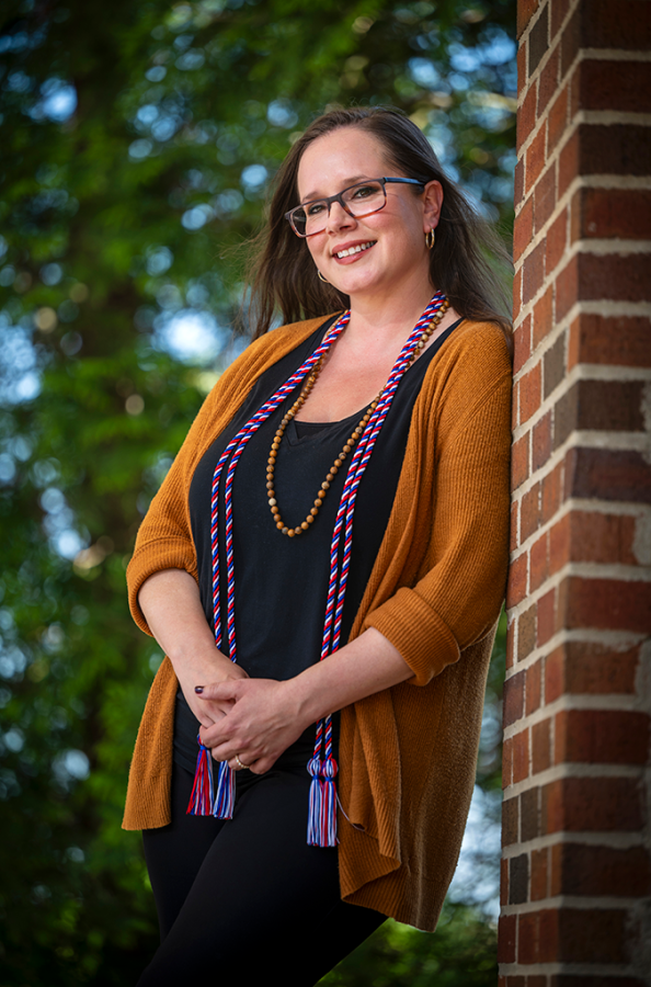 A woman wearing glasses and graduation cords smiles at the camera.