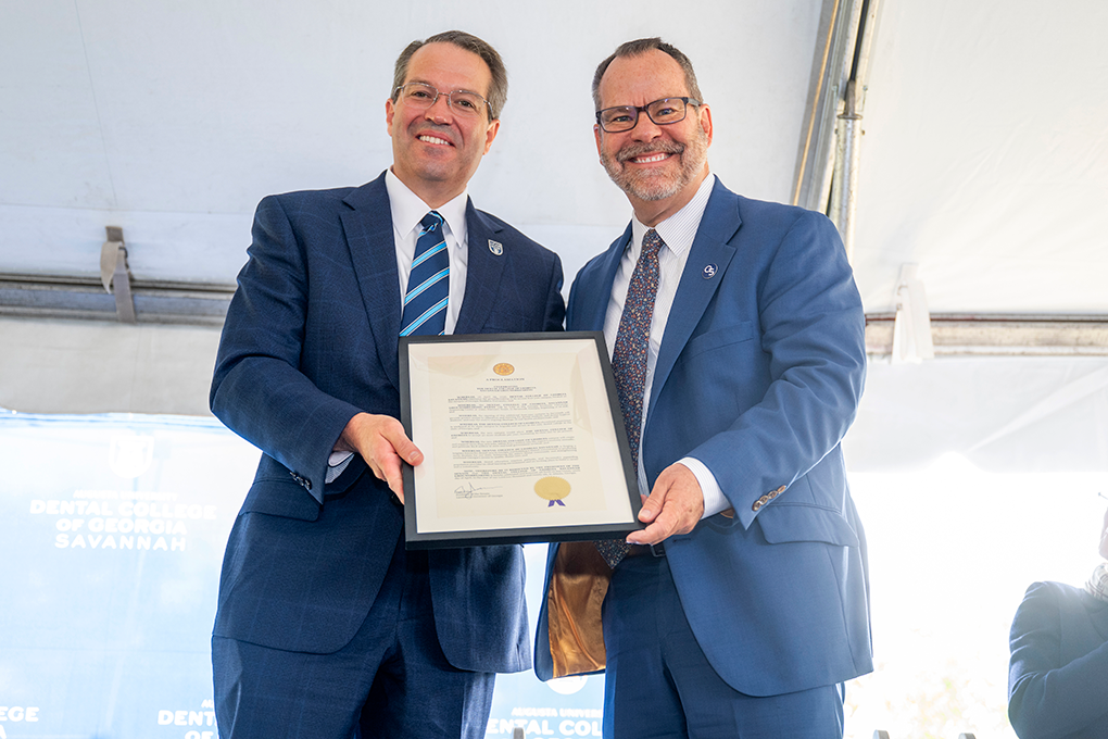 Two men wearing suits hold a plaque and smile at the camera.