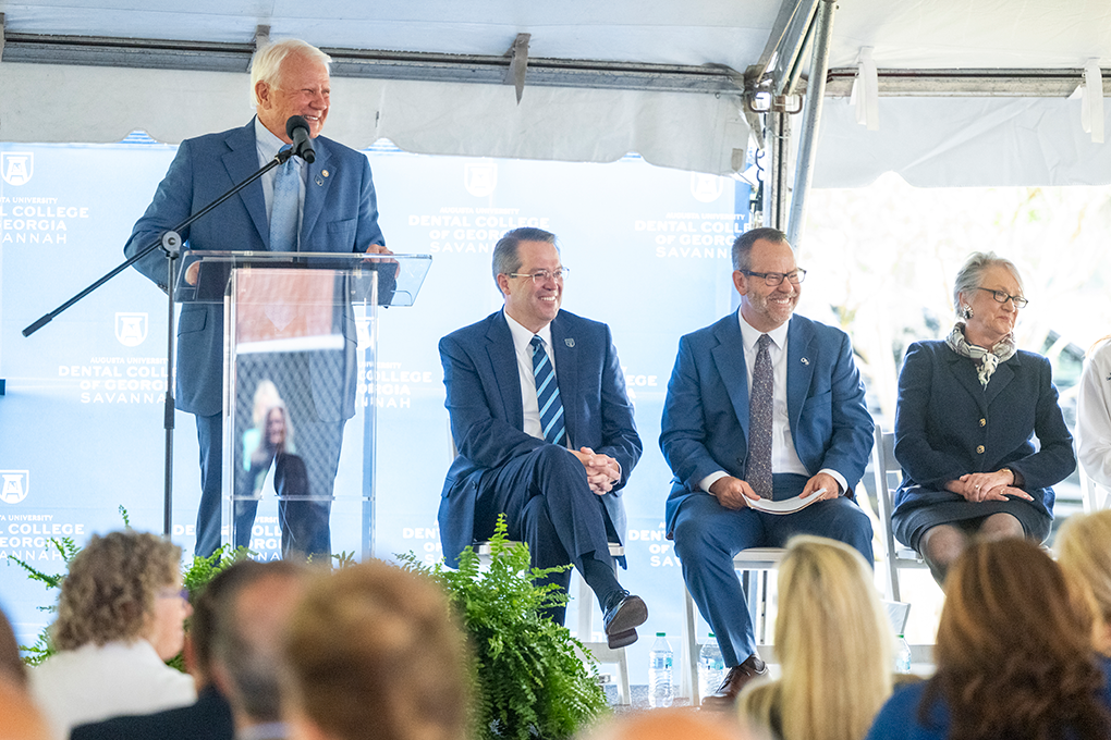 A man speaks at a podium while two men and a woman sit in chairs beside him.