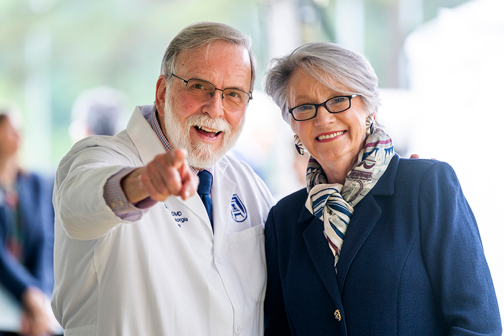 A man wearing a lab coat points and smiles at the camera while a woman smiles beside him.
