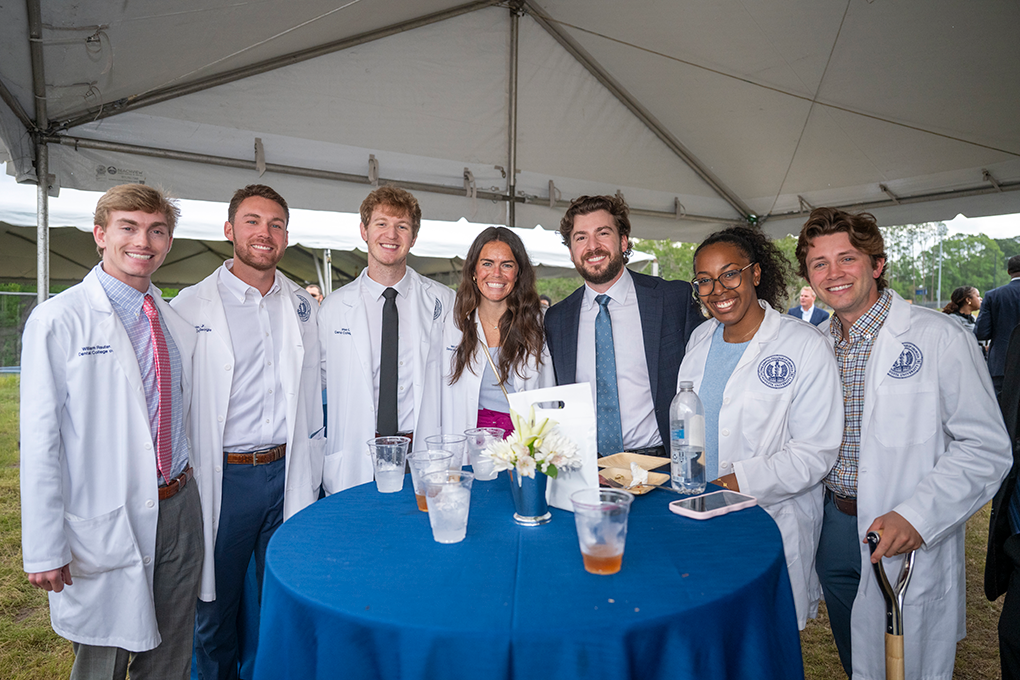 A group of dental students wearing lab coats stands around a table and smiles.
