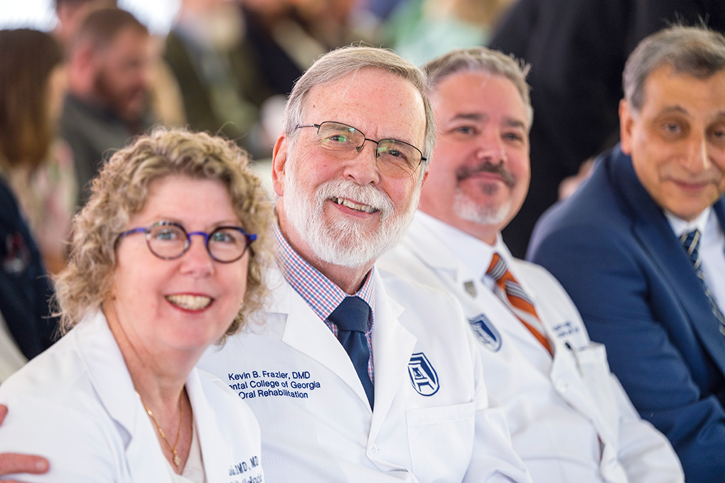 Four people sitting in a crowd smile at the camera. Three are wearing lab coats and the other is wearing a suit.