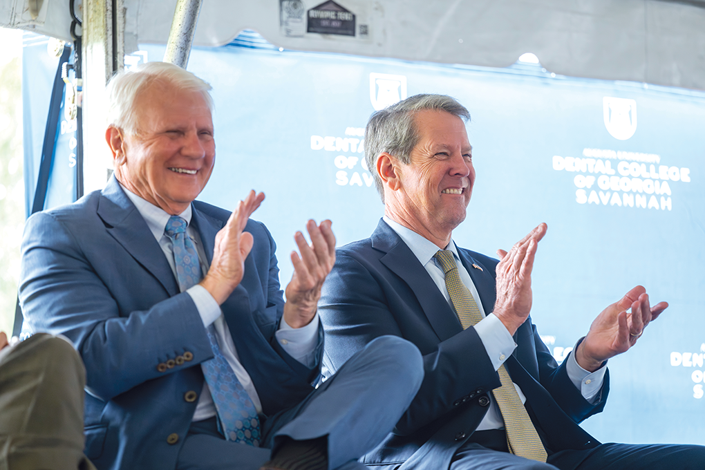 Two men wearing suits sit on a stage and clap.