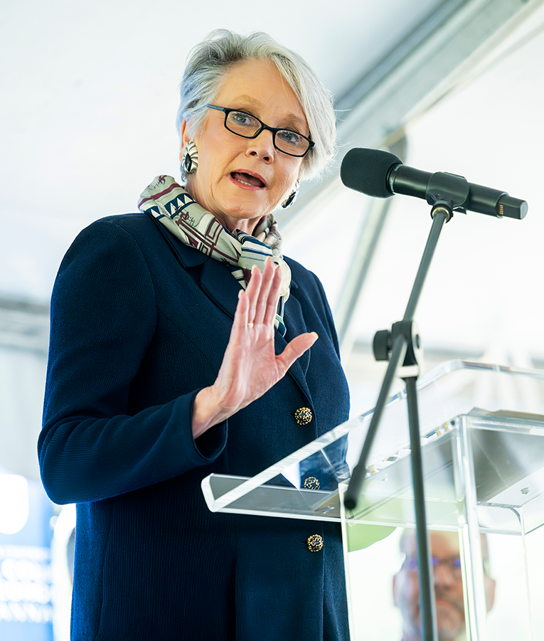A woman in a suit stands behind a podium and delivers a speech.