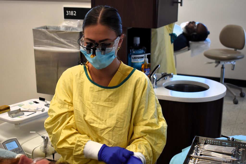 A young woman in a medical scrub, mask, gloves, and dental hygiene glasses sits with a patient