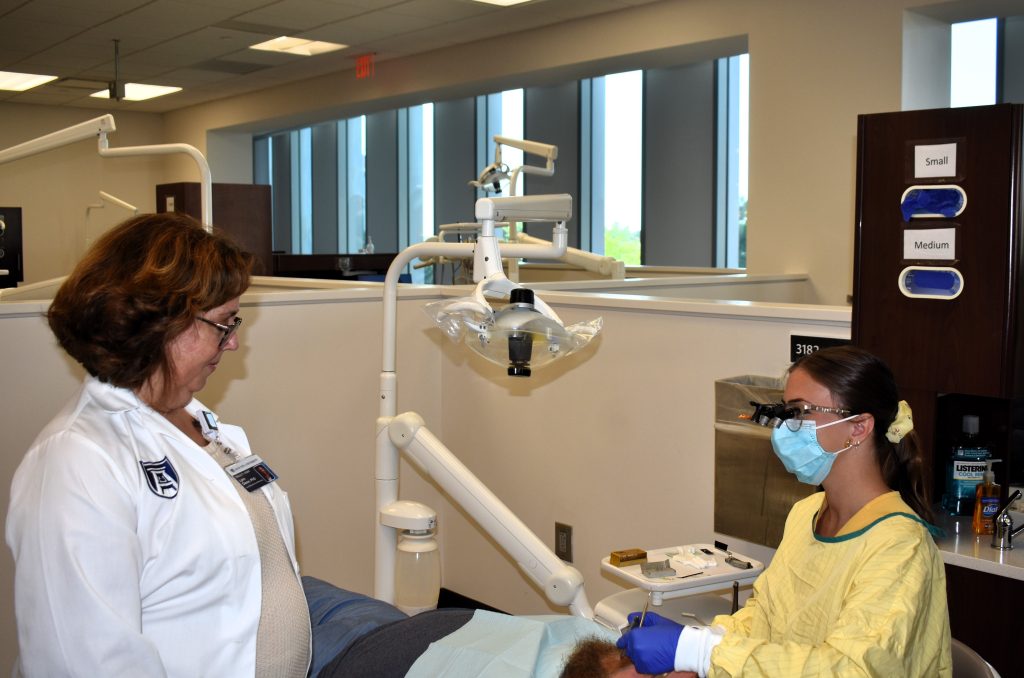 A woman in a white coat stands as a dental hygiene student works on a patient