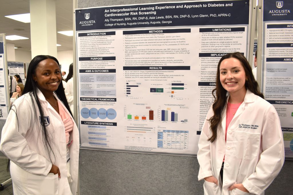 Two DNP students stand on either side of a scientific poster. They are wearing white coats and smiling at the camera