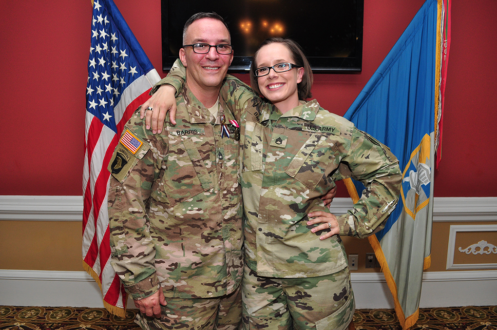 A man and a woman in army uniforms smiling and posing in front of United States flags.