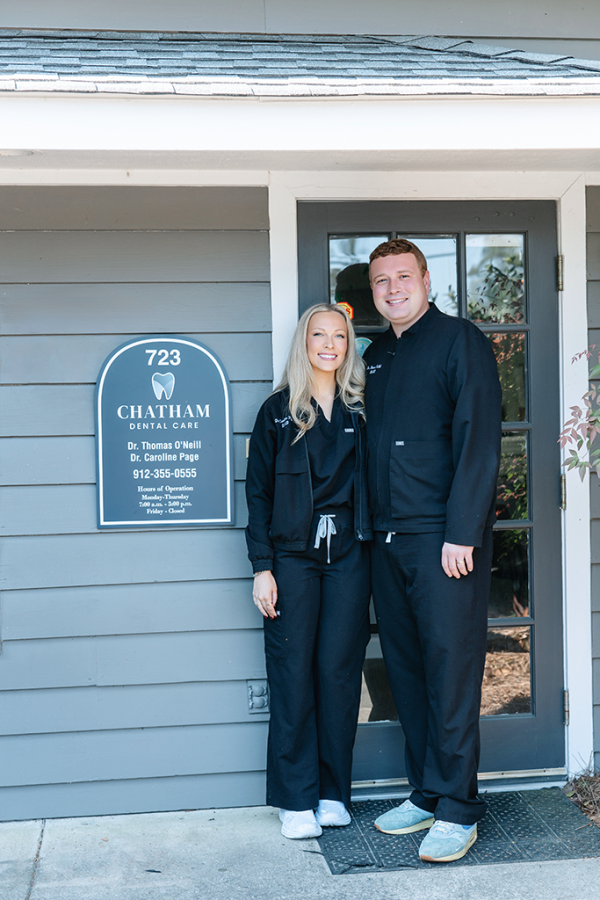 A man and a woman wearing scrubs stand in front of a dental office and smile at the camera.