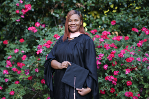 Young woman clothed in graduate regalia standing in front of flower bushes.