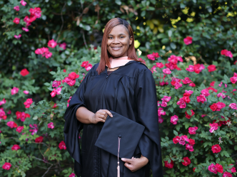 Young woman clothed in graduate regalia standing in front of flower bushes.