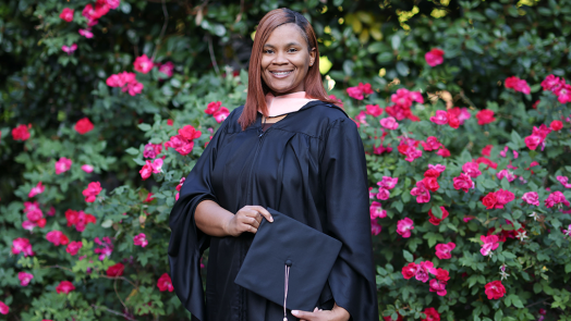 Young woman clothed in graduate regalia standing in front of flower bushes.