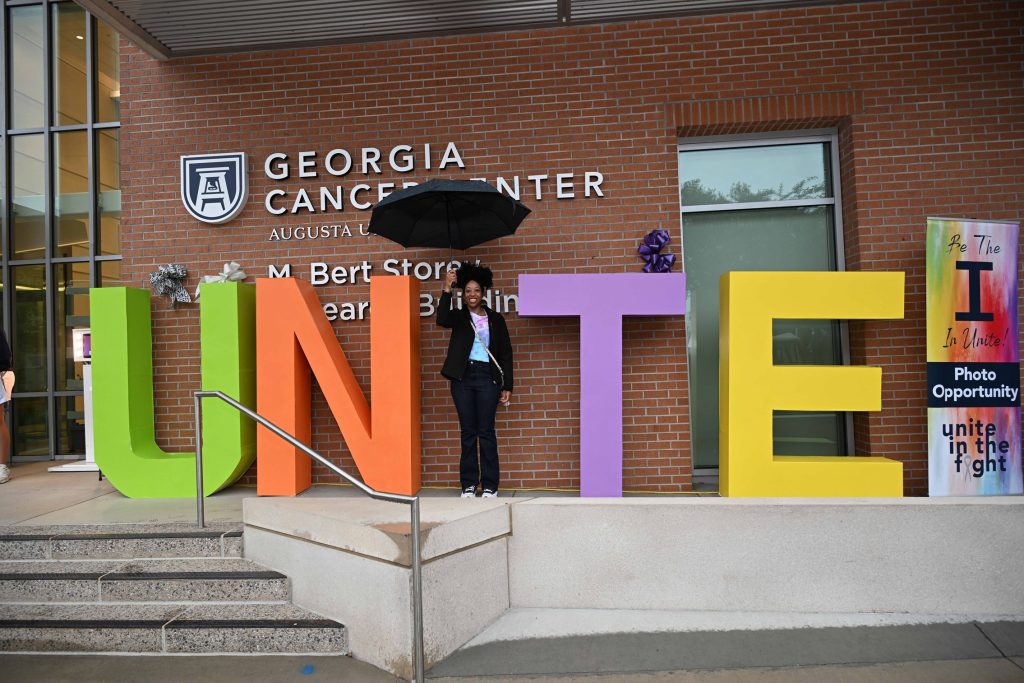 In this photo, you see a person standing and posing for a photo. The person is the "I" in the word UNITE. The letters U, N, T, and E are painted bright colors. People are encouraged to be the "I" to spell out unite.