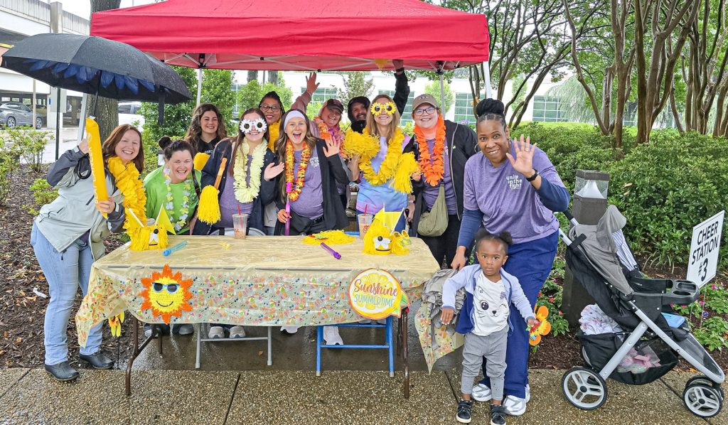 In this photo, you see people standing under a tent posing for a photo. These people are part of the Georgia Radiation Therapy Cheer Station.