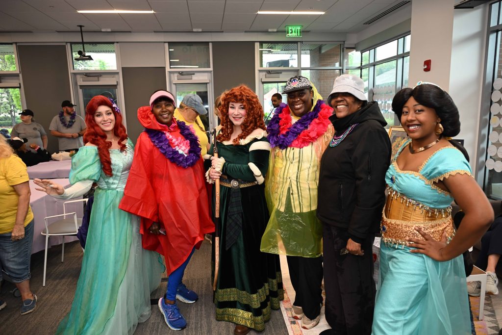 In this photo, you see people posing for a photo with a group of Disney princesses including Ariel, Jasmine, and Merida.