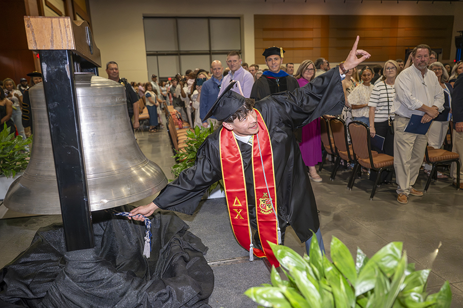 a male student in a graduation robe rings a silver bell and points to the sky 