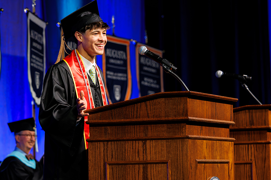male student in graduation robe smiling at a podium