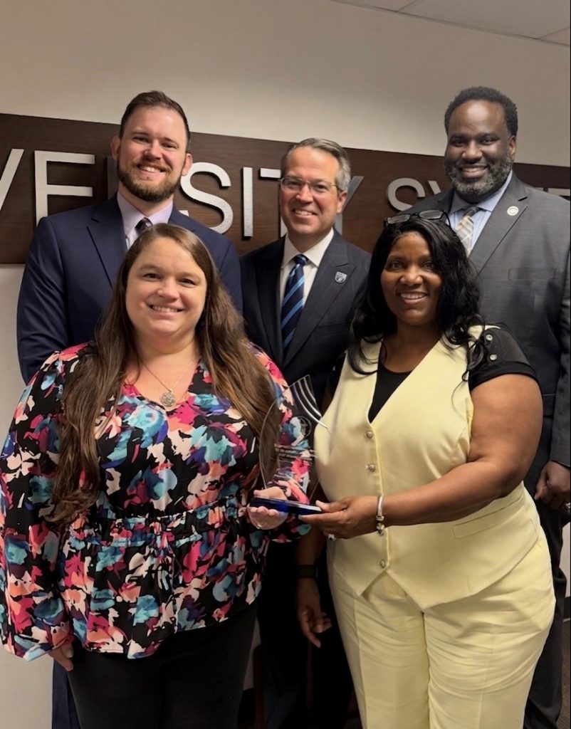 Three men and two women, who are holding an award, smile for a photo while indoors.