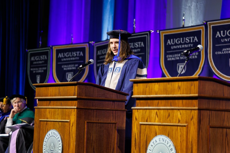 a male student in a blue and gray graduation gown speaks at a podium