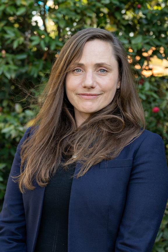 A woman in a blazer in front of greenery.