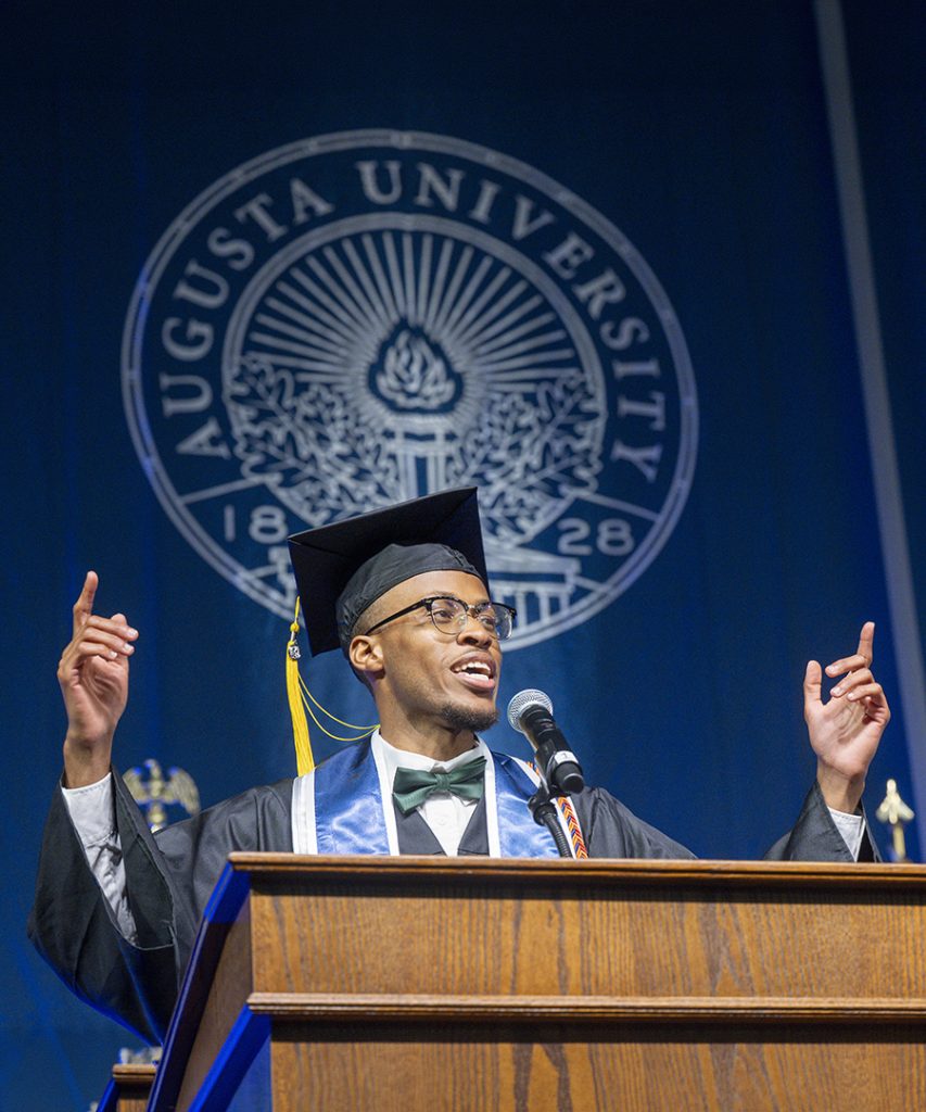 male student in graduation robe speaks at podium with his hands raised.