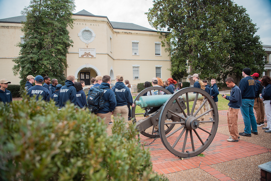 A group of students gathered around a historic yellow building with a vintage cannon in the foreground